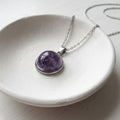 A silver chain necklace with a round purple amethyst cabochon pendant, displayed on a textured white ceramic dish.