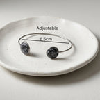An adjustable silver-tone cuff bracelet with two round dark gray and white marbled stone end caps, displayed on a small white ceramic dish with a measurement overlay indicating 6.5cm.