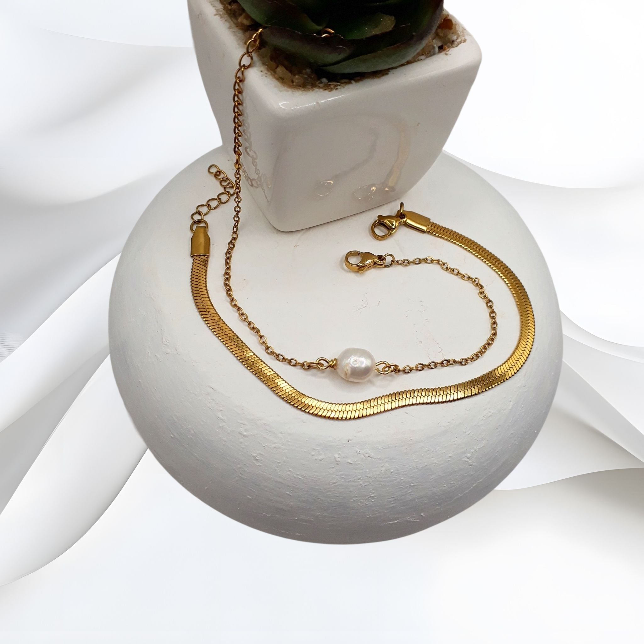 Gold-plated stainless steel double-layered bracelet with a solitaire white freshwater pearl and herringbone chain, displayed on a white ceramic surface.