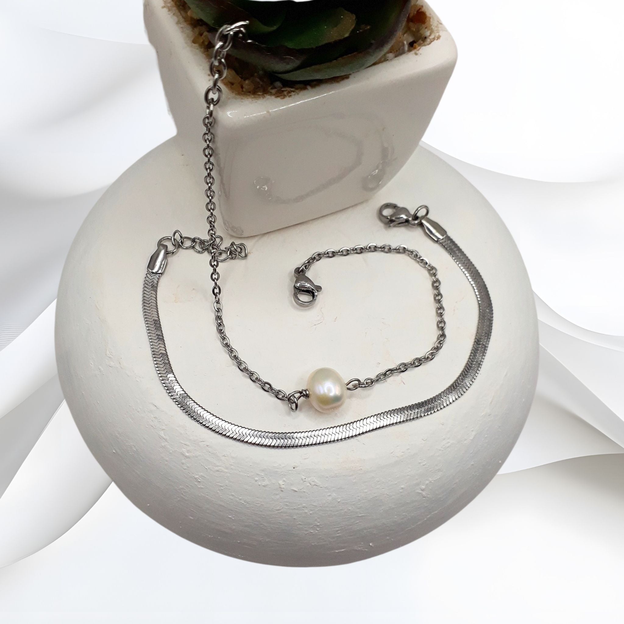 Stainless steel double-layered bracelet featuring a solitaire white freshwater pearl and a herringbone chain, displayed on a white ceramic surface.