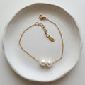A delicate gold chain bracelet with two white freshwater pearls, displayed on a textured white ceramic dish.