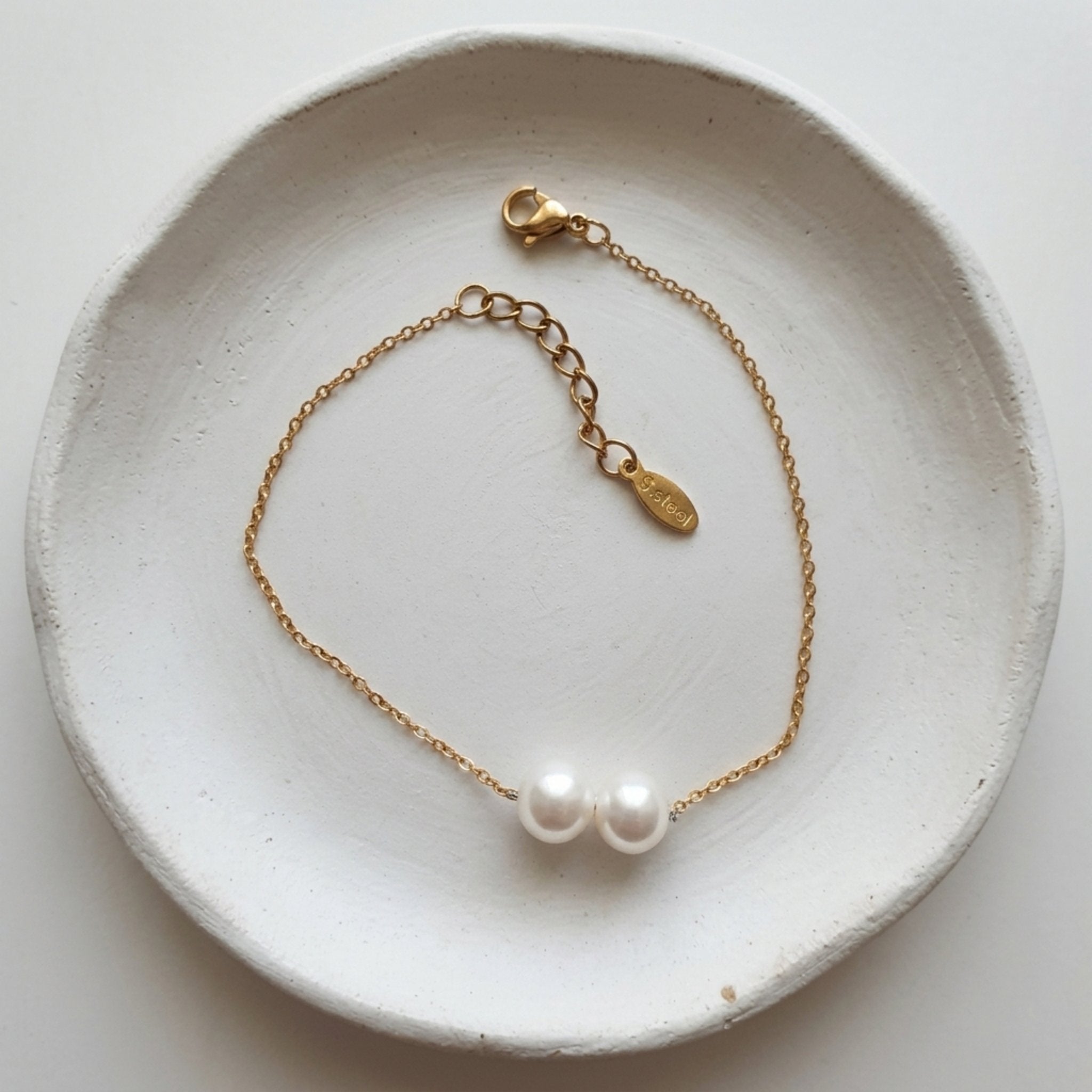 A delicate gold chain bracelet with two white freshwater pearls, displayed on a textured white ceramic dish.