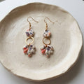 A pair of handmade dangle earrings with gold-tone hooks and rings, featuring small round beads and irregular clay charms in a white base with purple and pink floral patterns, displayed on a textured beige ceramic plate.