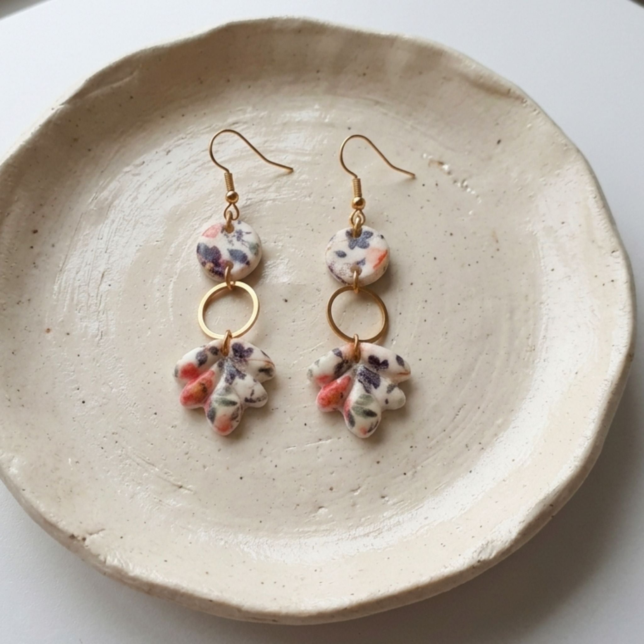 A pair of handmade dangle earrings with gold-tone hooks and rings, featuring small round beads and irregular clay charms in a white base with purple and pink floral patterns, displayed on a textured beige ceramic plate.