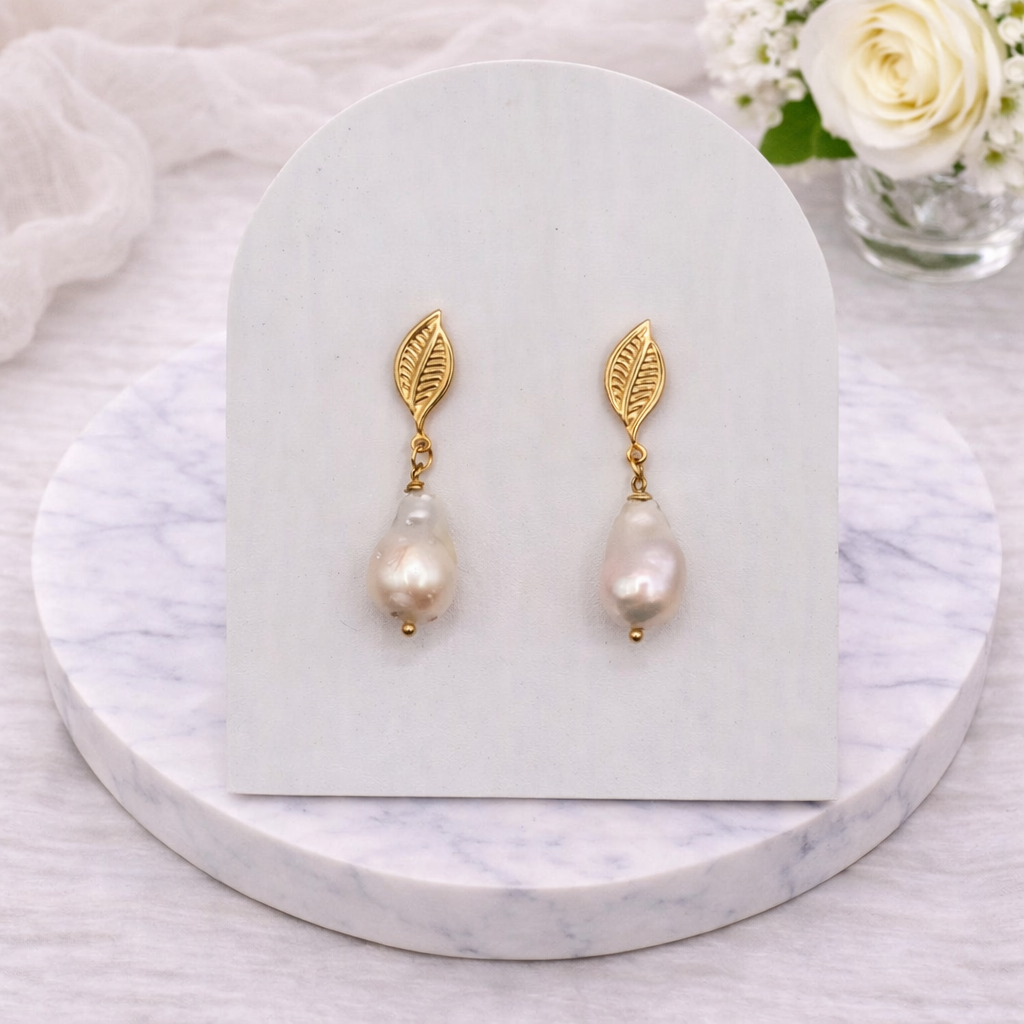 Gold leaf earrings with baroque pearls on a white card, placed on a marble surface with flowers in the background.