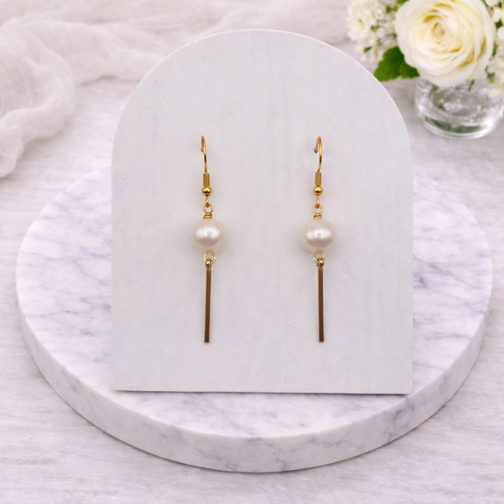 Pair of gold plated stainless steel pearl drop earrings on a white card with a marble base.