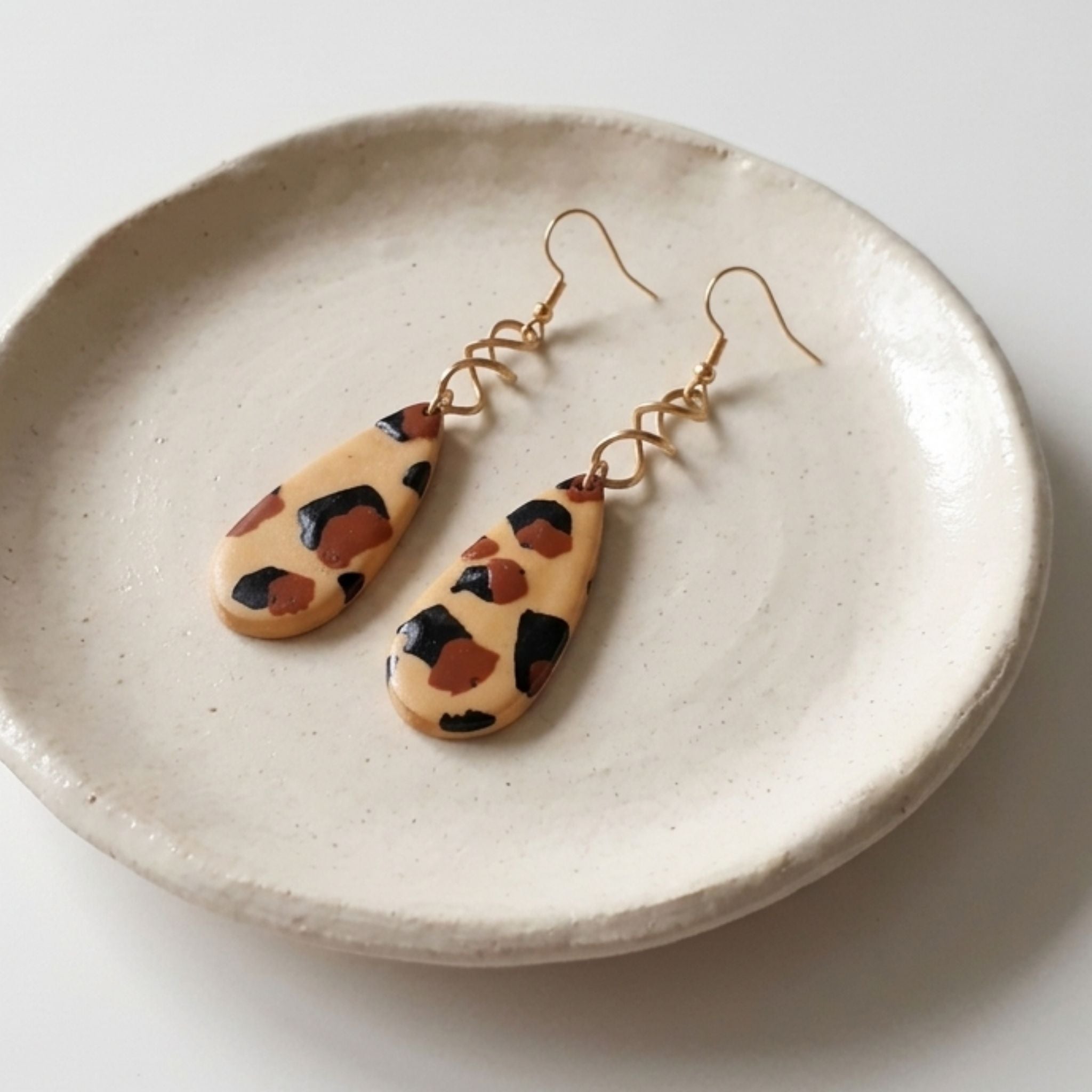 A pair of teardrop-shaped drop earrings with a beige, brown, and black leopard print pattern, hanging from gold-colored wire hooks on a textured cream ceramic dish.