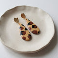 A pair of teardrop-shaped drop earrings with a beige, brown, and black leopard print pattern, hanging from gold-toned leaf-shaped posts, displayed on a textured beige ceramic dish.