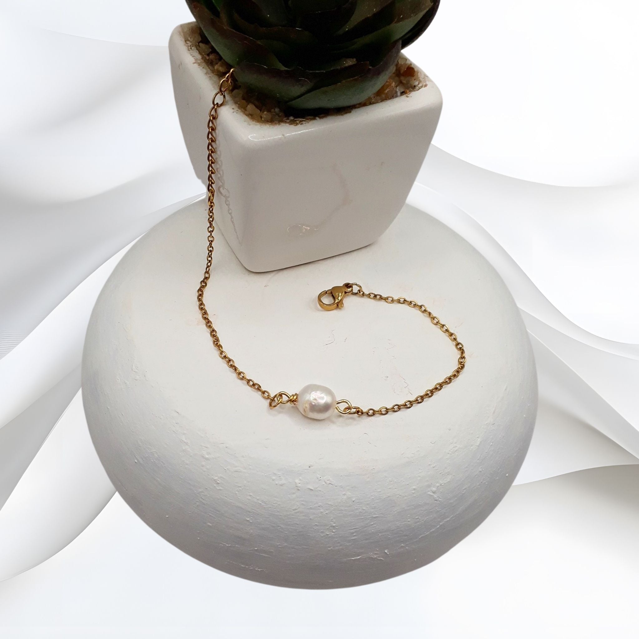 Gold-plated stainless steel bracelet with a single natural white freshwater pearl on a delicate cable chain, displayed on a white ceramic surface