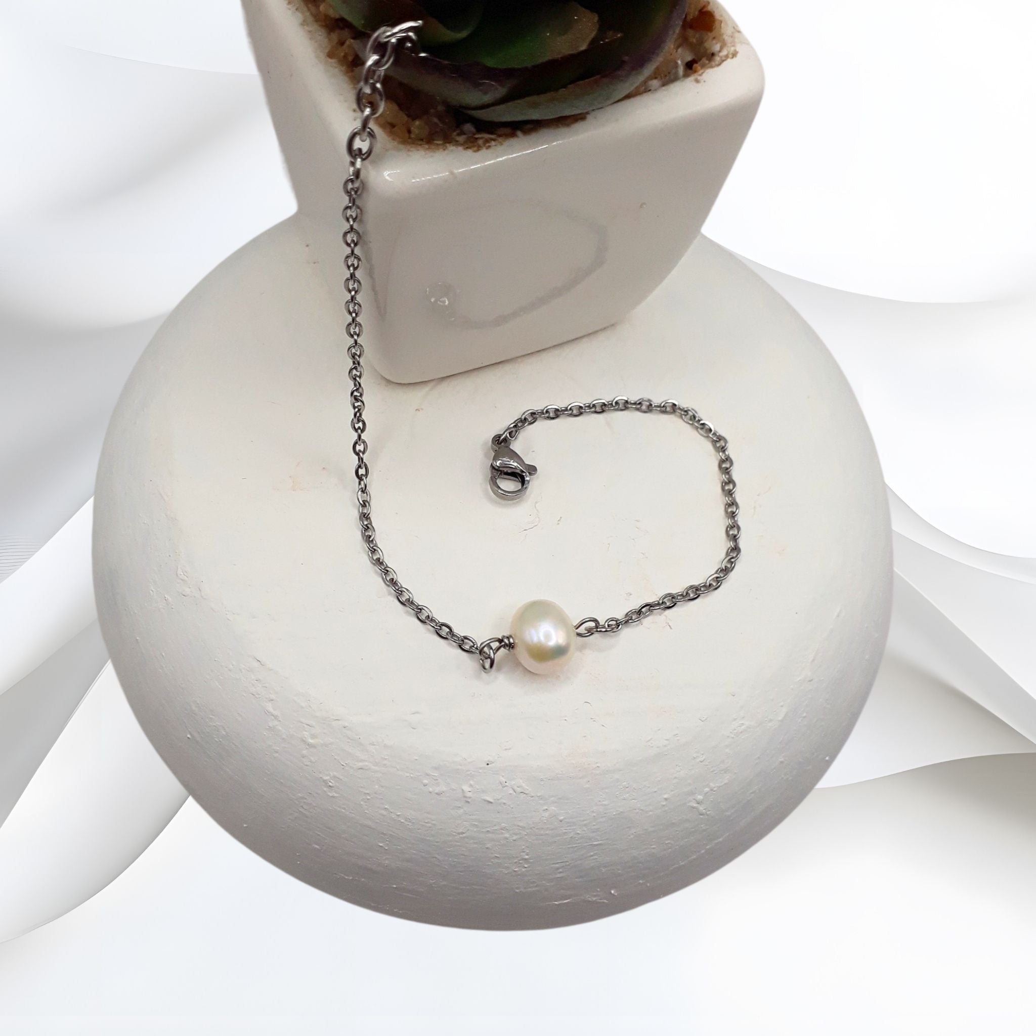 Stainless steel bracelet with a single white freshwater pearl on a delicate cable chain, displayed on a white ceramic surface.